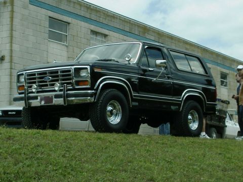1984 Ford Bronco - This black Ford Bronco is parked on a grassy incline with a building in the background. Another vehicle is visible to the left.