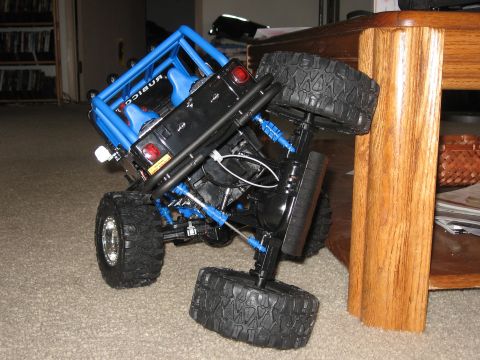 1990 Ford Bronco - A black remote control toy truck with a blue roll cage is tilted on a carpeted floor next to a wooden table.