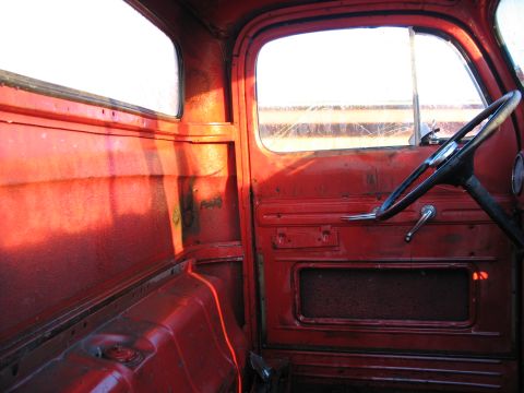 1952 Ford Pickup - The interior of a red truck cab shows a steering wheel and door panel with visible wear and tear.
