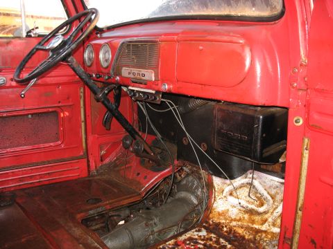 1952 Ford Pickup - The interior of a red 1952 Ford pickup shows its dashboard, steering wheel, and floor shifter, with visible rust on the floor.