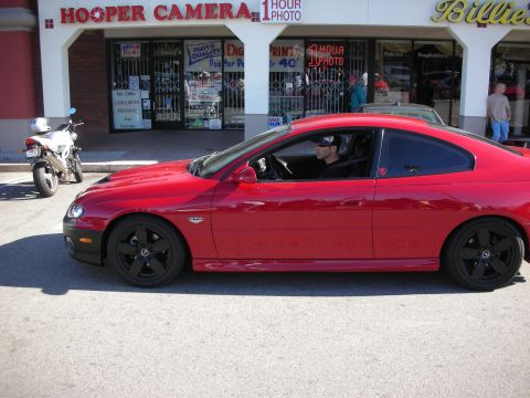 1998 Ford Crown Victoria - red two-door coupe with black wheels sits in profile on a street in front of a Hooper Camera photo shop and other storefronts.