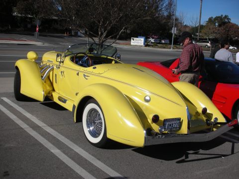 1998 Ford Crown Victoria - yellow boattail-style open roadster with chrome side exhausts, whitewall wire wheels and a red car beside it at a parking-lot meet.