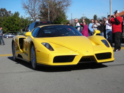 1998 Ford Crown Victoria - yellow Ferrari Enzo supercar rolls past a crowd of spectators at an outdoor car gathering on a sunny day.