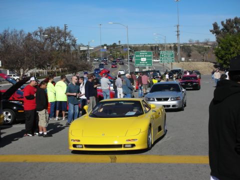 1998 Ford Crown Victoria - low yellow wedge-shaped exotic sports car faces the camera surrounded by spectators and other cars at a parking-lot meet.