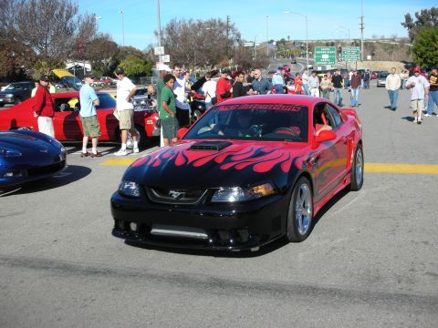 1998 Ford Crown Victoria - black Ford Mustang with red flame graphics and a hood scoop shown front three-quarter among crowds at an outdoor car meet.
