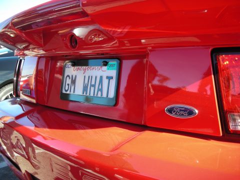 1998 Ford Crown Victoria - Close-up of a red car's rear showing a blue Ford oval emblem and a California 'GM WHAT' vanity plate dated 2007.