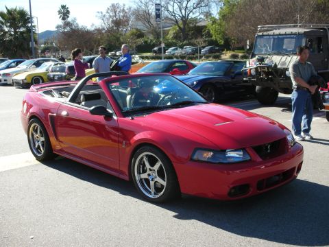 1998 Ford Crown Victoria - red Mustang Cobra convertible sits top-down with chrome wheels among other cars and spectators at a meet.