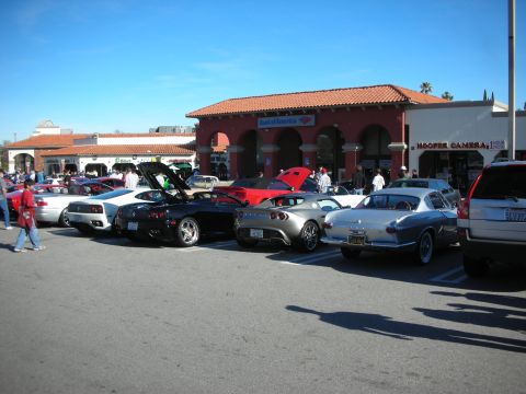1998 Ford Crown Victoria - lineup of sports cars, including exotics and a vintage coupe, parked before Spanish-style storefronts at a car meet.