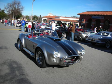1998 Ford Crown Victoria - silver Shelby Cobra replica roadster with black racing stripes rolls past spectators at an outdoor car meet.