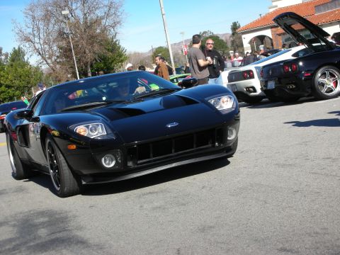 1998 Ford Crown Victoria - black Ford GT supercar rolls through a crowded outdoor car show with spectators and other sports cars in the background.