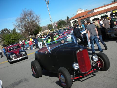 1998 Ford Crown Victoria - matte-black highboy hot rod roadster with red wheels cruises a packed car show lot, a maroon vintage coupe behind it.