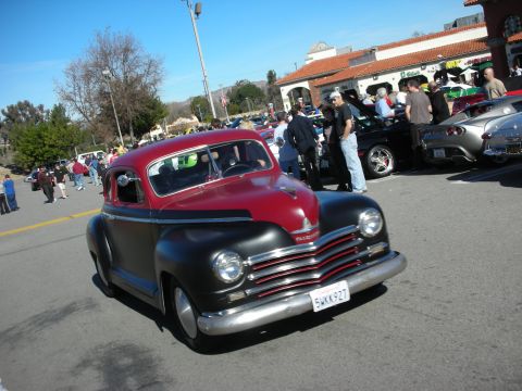1998 Ford Crown Victoria - red-and-black two-tone late-1940s Plymouth coupe street rod drives through a sunny car show parking lot.
