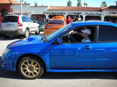 1998 Ford Crown Victoria - blue Subaru Impreza WRX STI with gold wheels idles at a car show, its driver in a white cap visible through the window.
