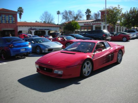 1998 Ford Crown Victoria - red Ferrari Testarossa with its signature side strakes drives through a car show parking lot lined with other sports cars.
