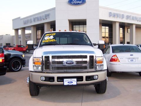 2000 Ford Excursion - A white Ford F-Series Super Duty truck is parked in front of a Ford dealership, with a white sedan to its right.