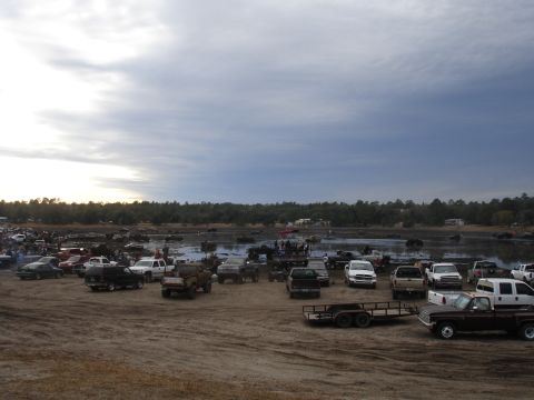 1989 Ford Bronco - A large gathering of trucks and SUVs are parked in a muddy field, with a body of water and trees in the background.