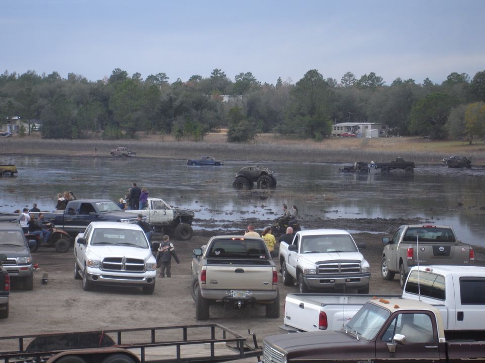 Muddy field with many trucks, ATVs, and a monster truck. Some vehicles are stuck in the mud