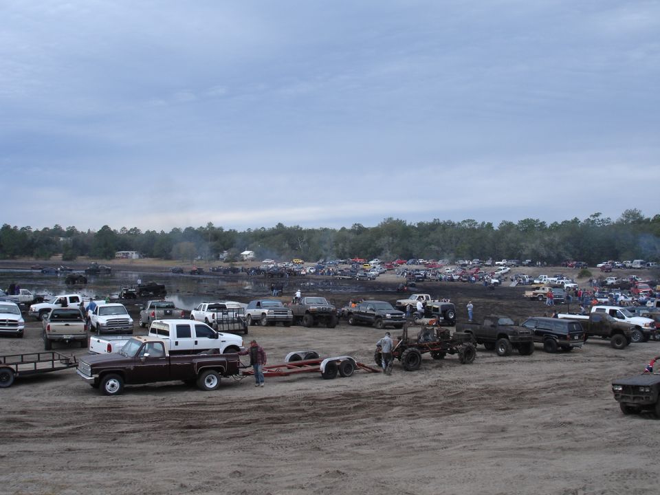 lots of trucks and people at a mud bog, some trucks are towing trailers