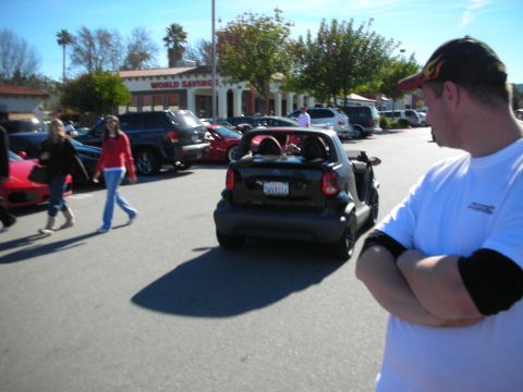 1998 Ford Crown Victoria - small black Smart Fortwo drives away through a busy car show parking lot as a man in a white shirt watches.