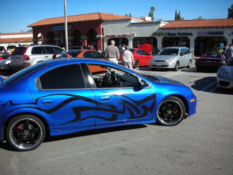 1998 Ford Crown Victoria - blue Dodge Neon sedan with black tribal-flame graphics and black wheels sits in a strip-mall car show lot.