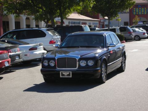 1998 Ford Crown Victoria - black Bentley Arnage luxury sedan with its quad headlights sits parked in a tree-lined shopping center parking lot.
