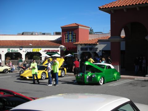 1998 Ford Crown Victoria - yellow Lamborghini and a lowered green custom hot rod truck are displayed by attendants outside a Subway and car-wash strip mall.