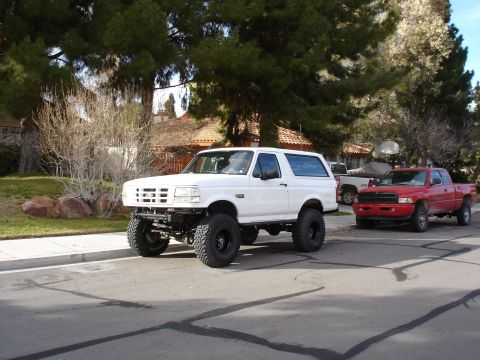 1996 Ford Bronco - A white Ford Bronco with large tires and a lifted suspension is parked on a residential street next to a red Dodge Ram pickup truck.