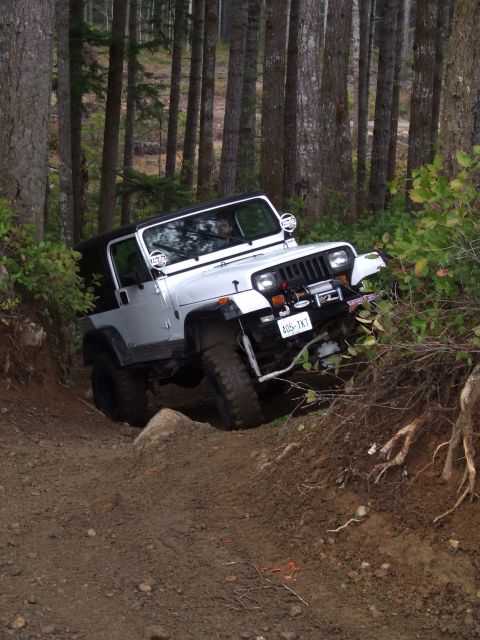 1991 Jeep Wrangler - A white 1991 Jeep Wrangler tackles a challenging off-road course through a wooded area.