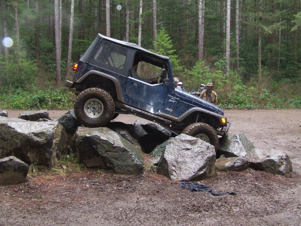 Dark blue 1991 Jeep Wrangler climbing over large rocks on a dirt trail, trees in background