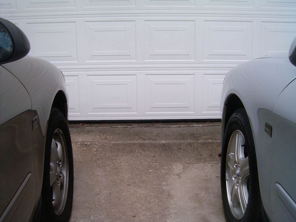 Two cars parked in front of a white garage door. The left car is dark, the right car is silver. Both show front wheel and fender