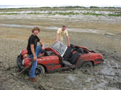 1974 Ford F-100 - Two men stand by a red convertible that is stuck in thick mud. The car appears to be a convertible with the top down.