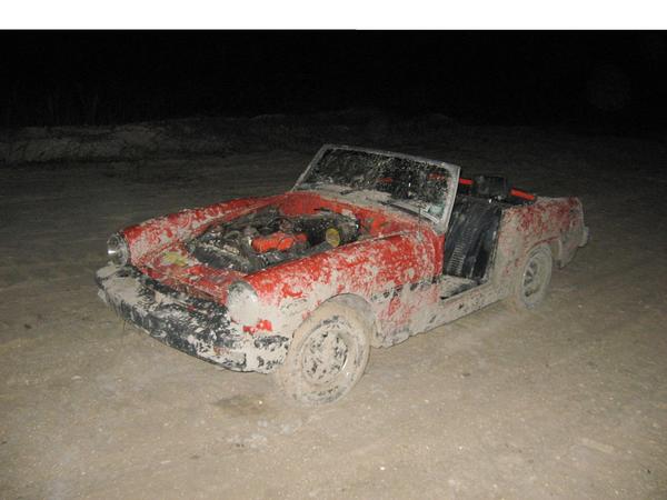 Mud-covered red convertible with hood open, showing engine. Driver's side door is missing
