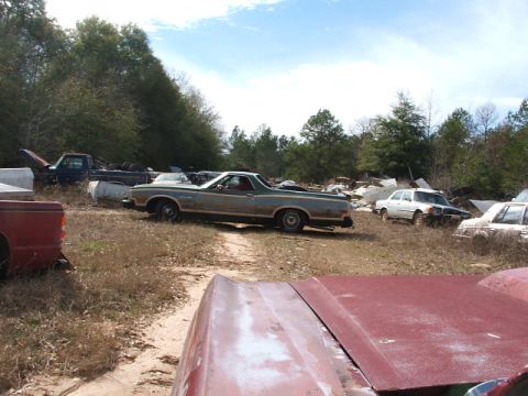 1966 Ford Ranchero - A weathered 1972 Ford Ranchero sits among other vehicles in a junkyard under a partly cloudy sky.