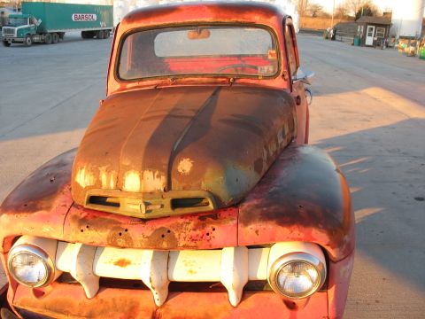 1952 Ford Pickup - A weathered red pickup truck sits in a lot, its grille showing distinct white bars.