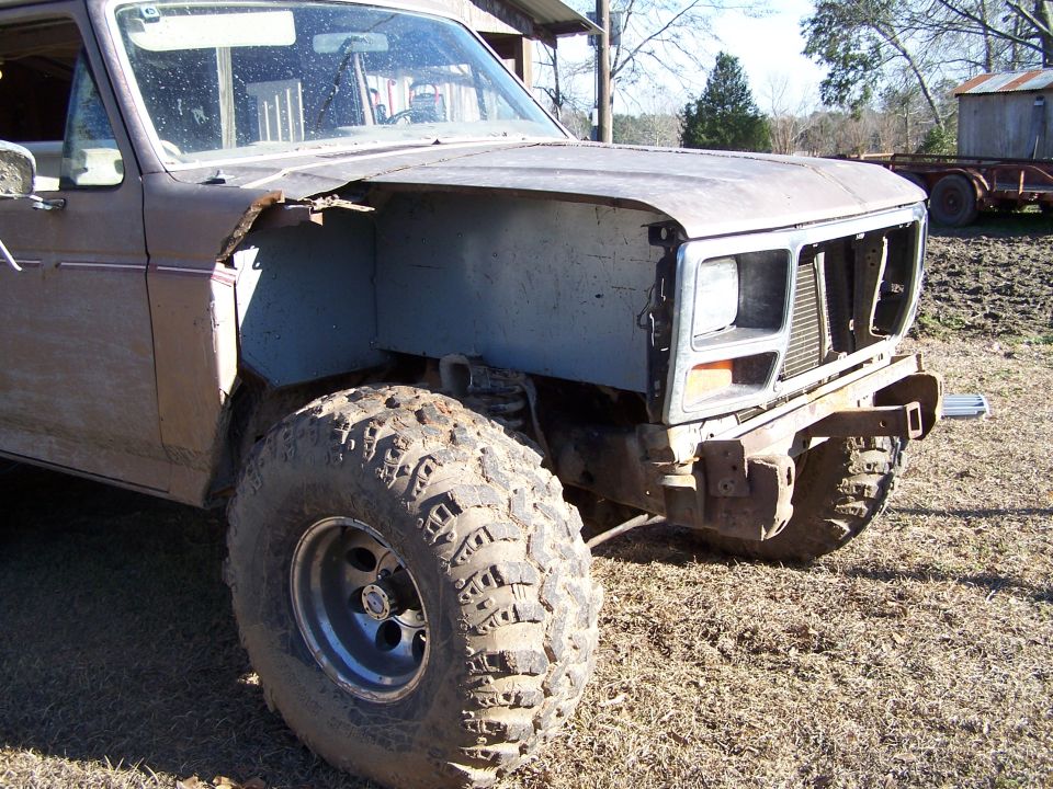 Brown Ford Bronco with missing fender and grille, large off-road tires, and a rusty bumper