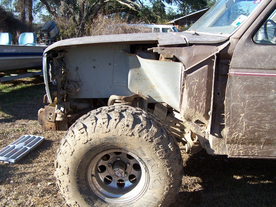 Front wheel and fender of a muddy, partially disassembled truck. A boat is visible in the background