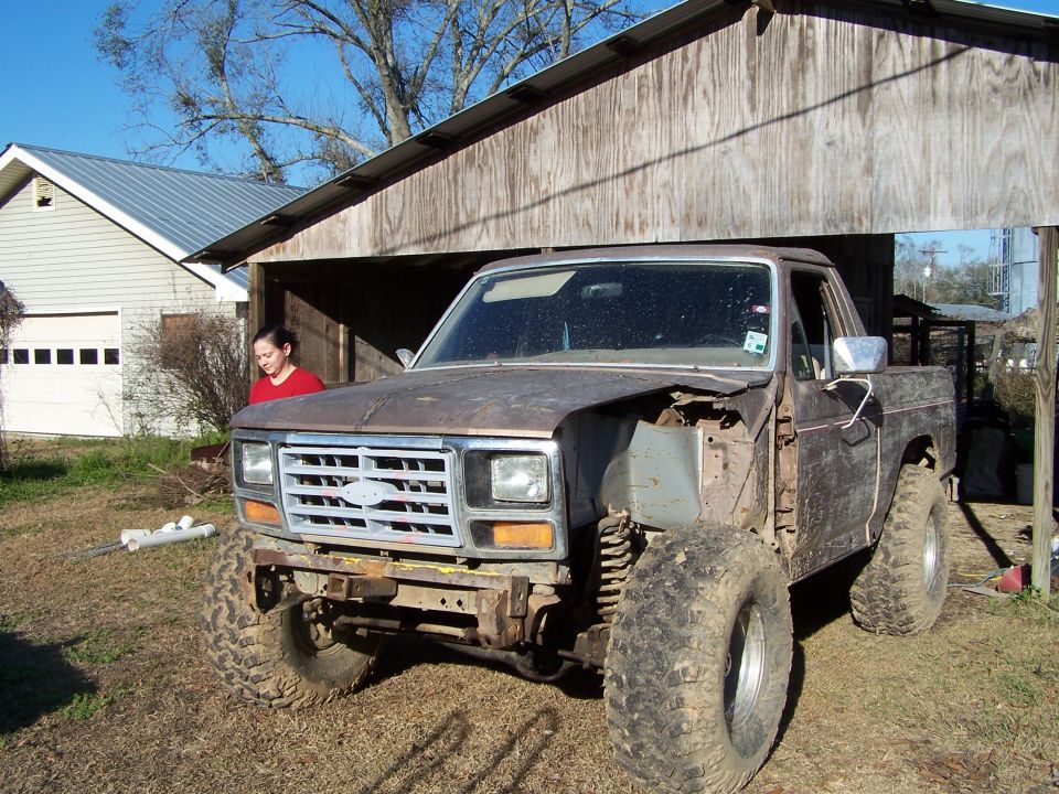 Brown Ford F-Series truck with missing fender, lifted suspension, and large tires parked under a wooden structure