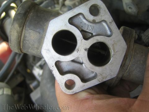 1990 Ford Bronco - hand holds a cast-metal engine part with two round ports and a figure-eight internal passage above an engine bay.