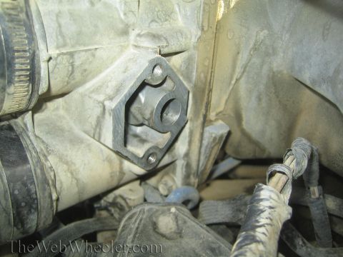 1990 Ford Bronco - diamond-shaped metal flange with bolt holes mounted against a dusty engine block with wiring nearby.