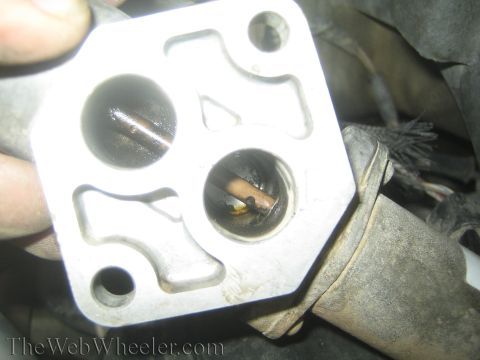 1990 Ford Bronco - hand holds a square metal flange with two ports, one showing a corroded brass fitting, over an engine area.