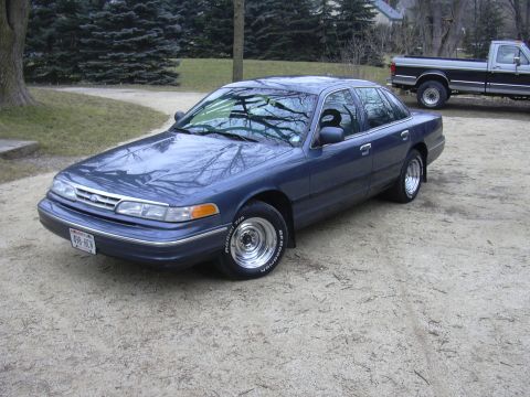 1996 Ford Crown Victoria - A blue 1996 Ford Crown Victoria sedan is parked on a gravel surface, with a pickup truck visible in the background.
