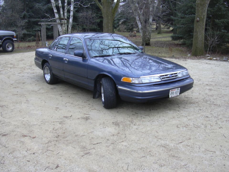 Blue 1995 Ford Crown Victoria parked on gravel, with a dark pickup truck to the left