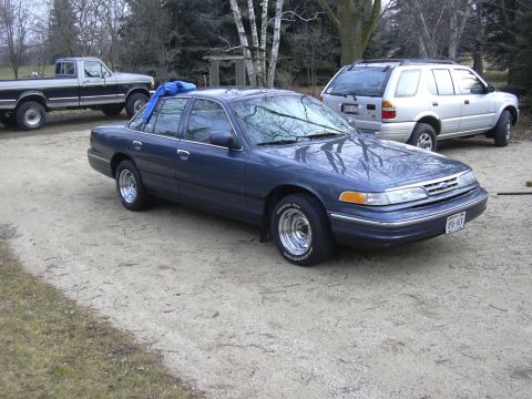 1996 Ford Crown Victoria - A blue 1996 Ford Crown Victoria sedan is parked in a gravel lot with other vehicles visible in the background.