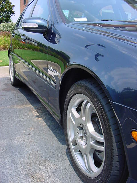 1984 BMW 3 Series - Close-up of a dark blue sedan's front fender and wheel, showing a V6 Kompressor badge.