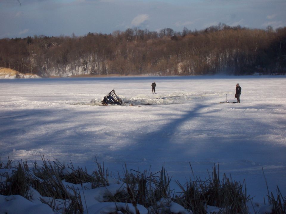 People standing on a frozen lake with broken ice and snowy tree-lined shore