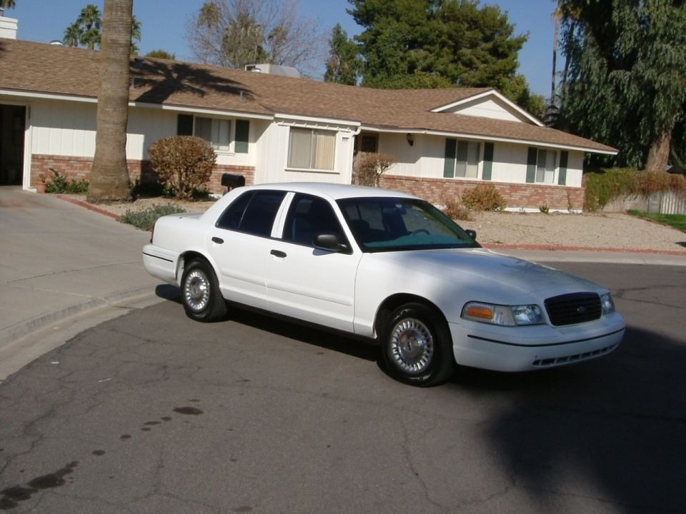 White Ford Crown Victoria sedan, front three-quarter view, parked on a street in front of a ranch house