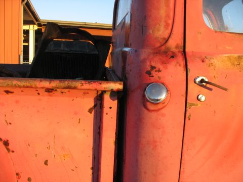 1952 Ford Pickup - The side of a weathered red pickup truck shows its door handle and a fuel cap, with a radiator visible in the truck bed.