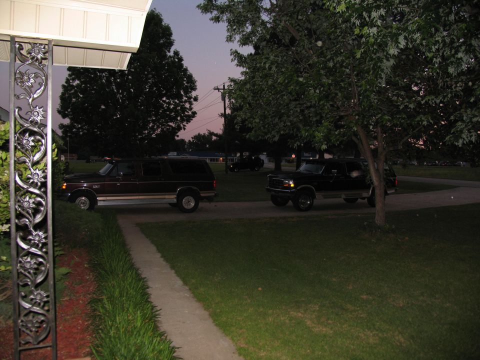 Two Ford Broncos parked on a driveway at dusk. The one on the left is dark red, the one on the right is black