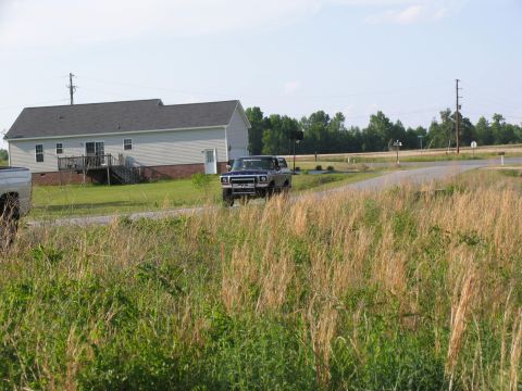 1995 Ford C-350 (Centurion Conversions) - A blue pickup truck drives down a road past a house and tall grass.