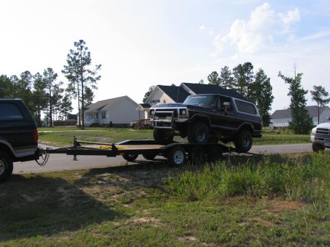 1995 Ford C-350 (Centurion Conversions) - A dark SUV tows a trailer carrying a black Ford Bronco past suburban houses.